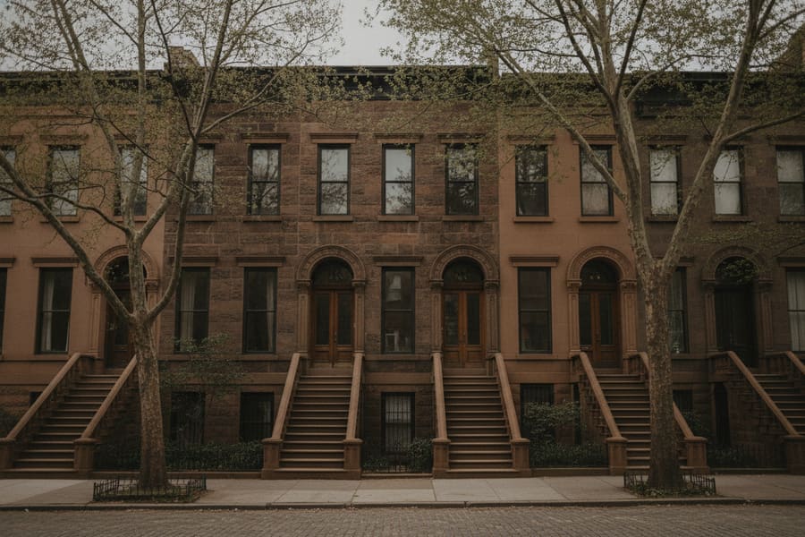 A row of Brooklyn brownstones with high stoops and tall parlour windows on a tree lined street